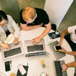 Team members meet around table with laptops